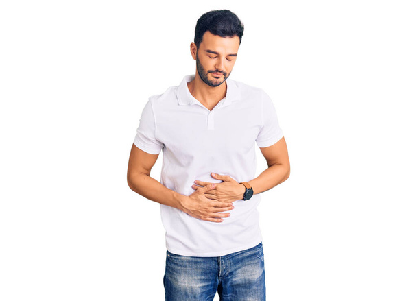 Jonge knappe Spaanse man die casual kleren draagt met de hand op de maag omdat indigestie, pijnlijke ziekte zich onwel voelt. pijnconcept.  - Foto, afbeelding