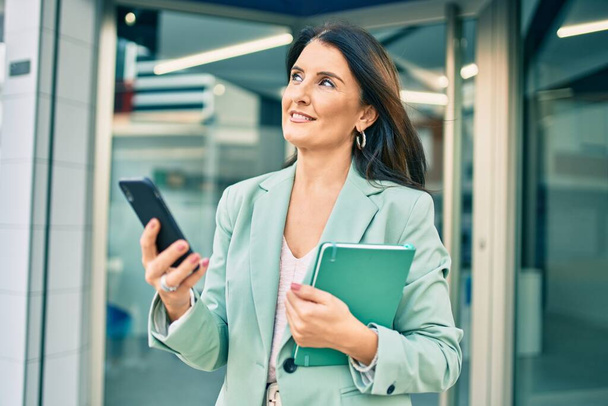 Mujer de negocios de confianza que usa un traje elegante de pie en la calle usando un teléfono inteligente con la cara alegre - Foto, imagen