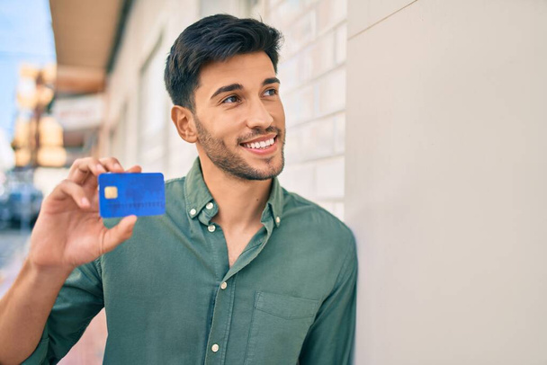 Jonge Latijnse man glimlachend met een kredietkaart op de muur in de stad.. - Foto, afbeelding
