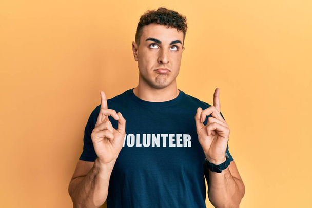 Hispanischer junger Mann im freiwilligen T-Shirt, das nach oben zeigt, sieht traurig und aufgebracht aus, gibt mit den Fingern die Richtung vor, unglücklich und deprimiert.  - Foto, Bild