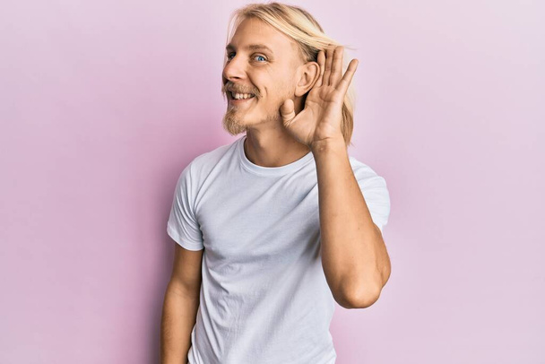 Ein kaukasischer junger Mann mit langen Haaren, der ein lässiges weißes T-Shirt trägt und lächelnd mit der Hand über dem Ohr Gerüchten oder Klatsch lauscht. Konzept für Gehörlosigkeit.  - Foto, Bild