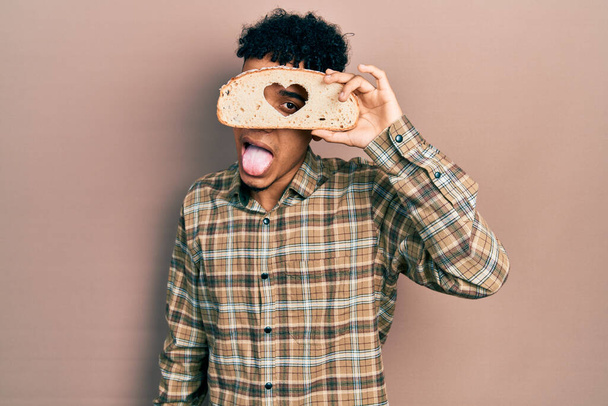 Young african american man holding bread loaf with heart shape sticking tongue out happy with funny expression.  - Фото, зображення