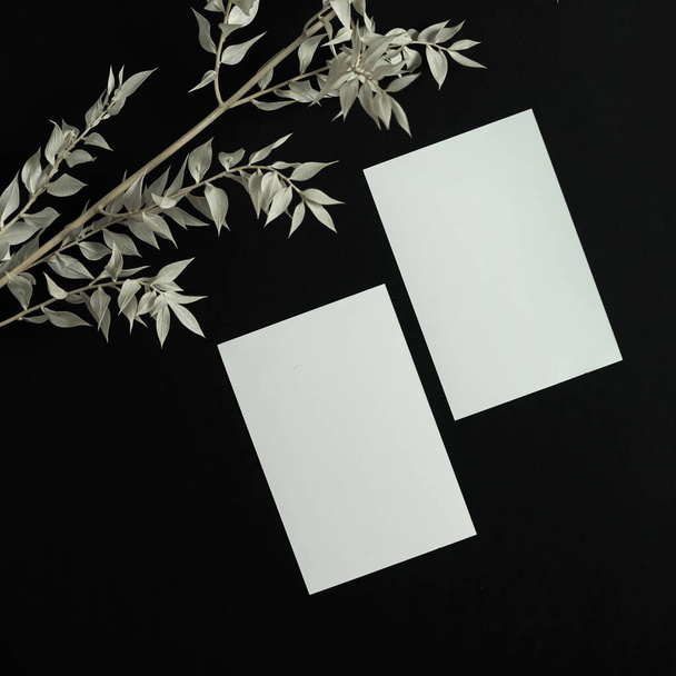 Leere Papierbögenkarte mit Kopierattrappe und trockenem Blumenzweig auf schwarzem Hintergrund. Minimale ästhetische Geschäftsmarken-Vorlage. Flache Lage, Draufsicht - Foto, Bild
