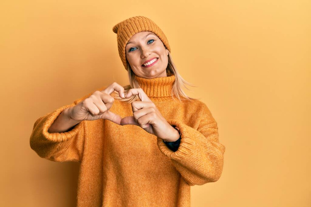 Middle age caucasian woman wearing wool winter sweater and hat smiling in love showing heart symbol and shape with hands. romantic concept.  - Фото, изображение