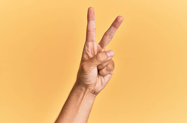 Paže a ruka bělocha přes žluté izolované pozadí počítání číslo 2 ukazující dva prsty, gestikulující vítězství a symbol vítěze  - Fotografie, Obrázek