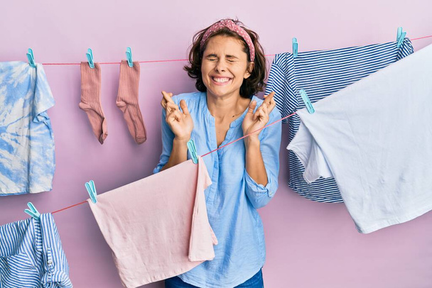 Νεαρή μελαχρινή γυναίκα που πλένει τα ρούχα της γύρω από σπάγκο κρέμεται χειρονομώντας σταυρωμένα δάχτυλα χαμογελώντας με την ελπίδα και τα μάτια κλειστά. τύχη και προληπτική έννοια.  - Φωτογραφία, εικόνα