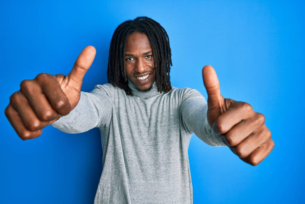 African american man with braids wearing turtleneck sweater approving doing positive gesture with hand, thumbs up smiling and happy for success. winner gesture.  - Photo, Image