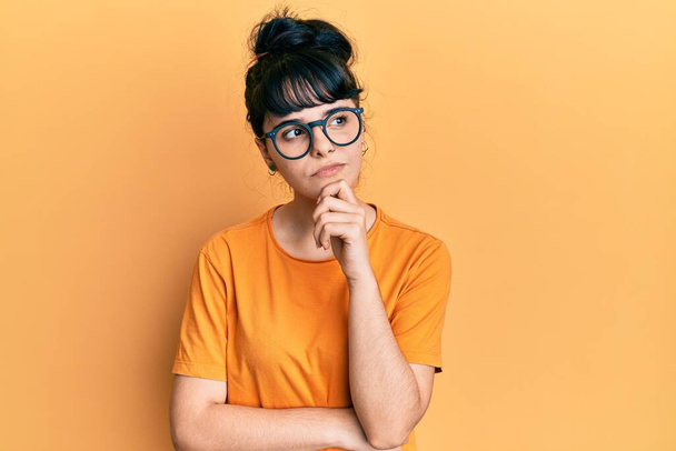 Young hispanic girl wearing casual clothes and glasses serious face thinking about question with hand on chin, thoughtful about confusing idea  - Photo, Image