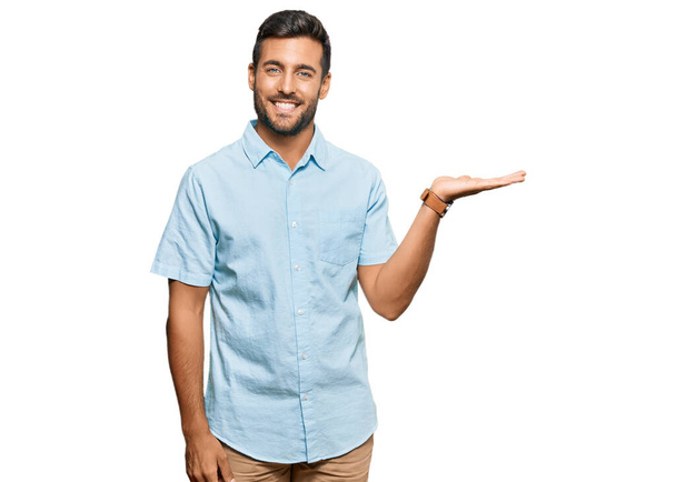 Hombre hispano guapo vistiendo ropa casual sonriendo alegre presentando y señalando con la palma de la mano mirando a la cámara.  - Foto, Imagen