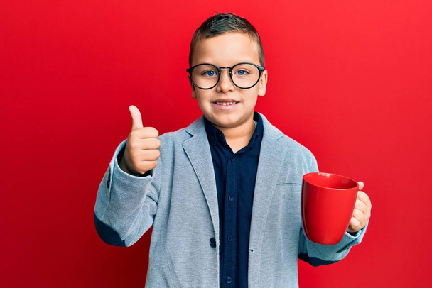 Malý chlapec nosí brýle pije z červeného hrnku usmívá šťastný a pozitivní, palec nahoru dělá vynikající a schvalovací znamení  - Fotografie, Obrázek