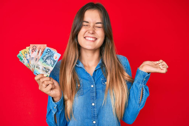 Schöne kaukasische Frau hält australische Dollars in der Hand und schreit stolz, feiert Sieg und Erfolg sehr aufgeregt mit erhobenem Arm  - Foto, Bild