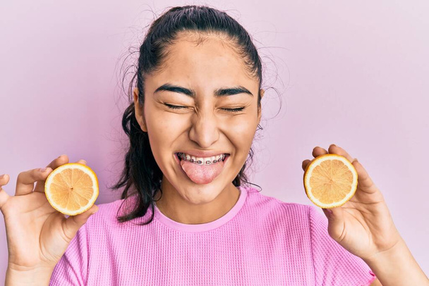 Hispanic teenager girl with dental braces holding lemon sticking tongue out happy with funny expression.  - Foto, imagen