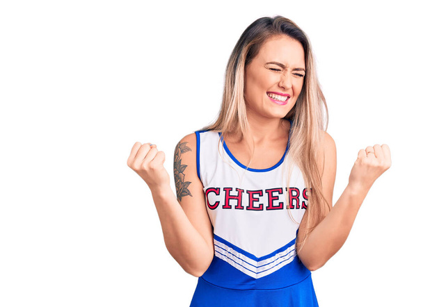 Giovane bella donna bionda indossa uniforme cheerleader molto felice ed eccitato facendo gesto vincitore con le braccia alzate, sorridente e urlando per il successo. concetto di celebrazione.  - Foto, immagini