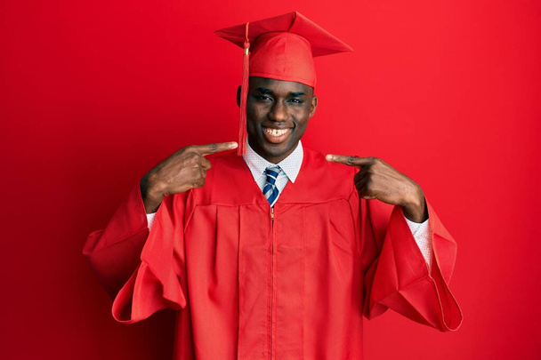 卒業キャップと儀式用のローブを着た若いアフリカ系アメリカ人の男性は、明るい表情と指の歯と口で指して笑っています。歯の健康の概念.  - 写真・画像