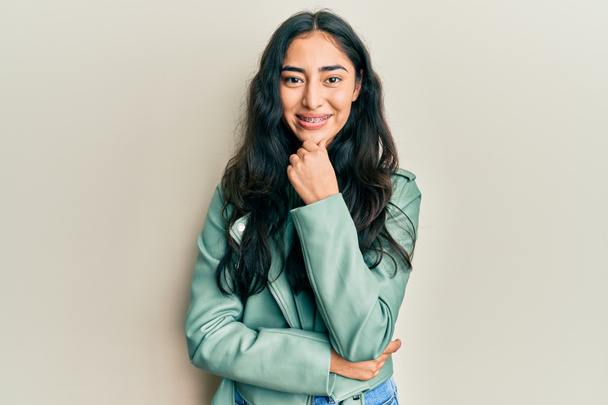 Hispanic teenager girl with dental braces wearing green leather jacket smiling looking confident at the camera with crossed arms and hand on chin. thinking positive.  - Fotó, kép