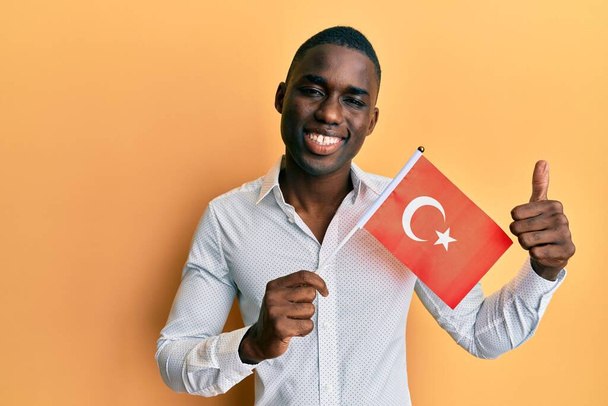 Young african american man holding turkey flag smiling happy and positive, thumb up doing excellent and approval sign  - Photo, Image