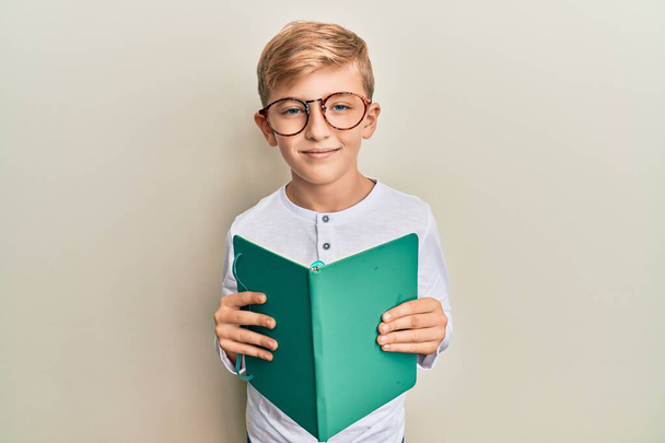 Pieni valkoihoinen poika, joka lukee kirjaa lasit päässään, rentoutuneena ja ilme kasvoillaan. yksinkertainen ja luonnollinen kameraan katsominen.  - Valokuva, kuva