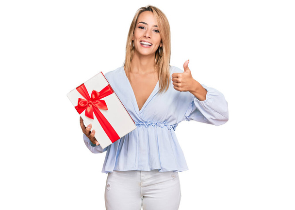 Όμορφη ξανθιά γυναίκα κρατώντας δώρο χαμογελώντας χαρούμενος και θετικός, αντίχειρας επάνω κάνει εξαιρετική και την έγκριση σημάδι  - Φωτογραφία, εικόνα
