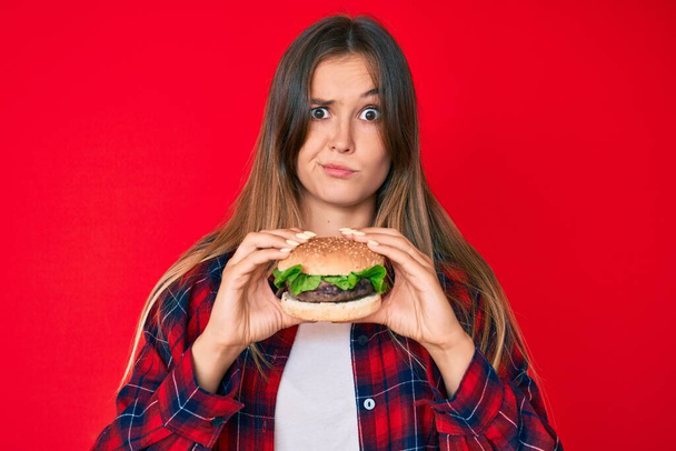 Schöne kaukasische Frau isst einen leckeren klassischen Burger skeptisch und nervös, Stirnrunzeln aufgeregt wegen des Problems. negative person.  - Foto, Bild