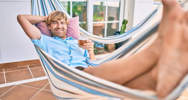 Hombre guapo de mediana edad en la terraza de su casa relajándose tumbado en una hamaca bebiendo una cerveza fresca - Foto, imagen