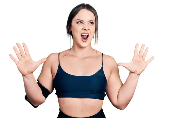 Mujer hispana joven usando ropa deportiva loca y loca gritando y gritando con expresión agresiva y los brazos levantados. concepto de frustración.  - Foto, Imagen