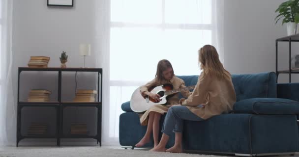 ελεύθερο χρόνο στο σπίτι, γυναίκα και κόρη κάνουν μουσική, κοριτσάκι μαθαίνει να παίζει κιθάρα, κάθεται στον καναπέ στο σαλόνι τα Σαββατοκύριακα - Πλάνα, βίντεο