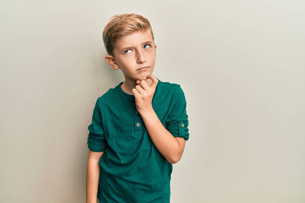 Kleiner kaukasischer Junge in lässiger Kleidung, der konzentriert mit dem Finger am Kinn über Zweifel nachdenkt und staunend nach oben blickt  - Foto, Bild