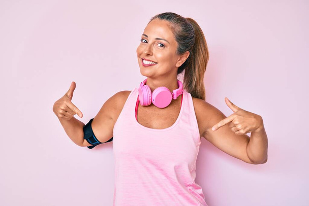 Mujer hispana de mediana edad vistiendo ropa de gimnasio y usando auriculares que parecen confiados con sonrisa en la cara, señalándose con los dedos orgullosos y felices.  - Foto, Imagen