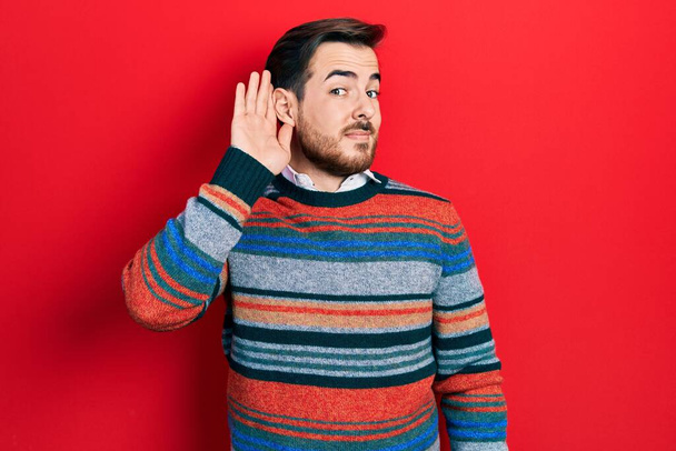 Красивий білий чоловік з бородою носить елегантний вовняний зимовий светр, посміхаючись рукою над вухом, слухаючи чутки або плітки. концепція глухоти
.  - Фото, зображення