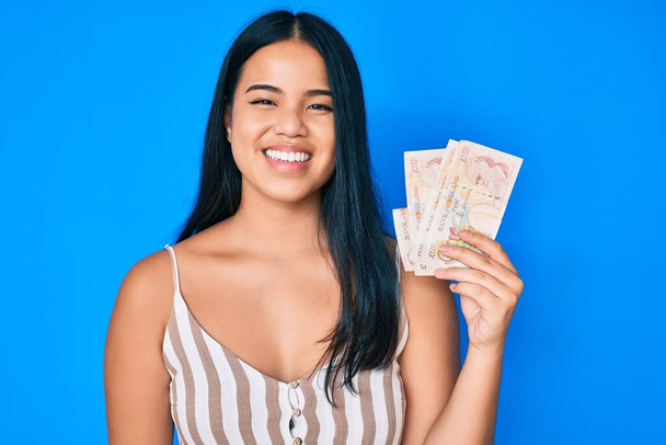 Young beautiful asian girl holding united kingdom pounds looking positive and happy standing and smiling with a confident smile showing teeth  - Photo, Image