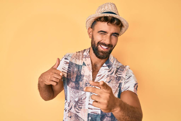 A fiatal spanyol férfi nyári kalapban ujjal mutogat a kamerára, boldog és vicces arccal. jó energia és rezgések.  - Fotó, kép