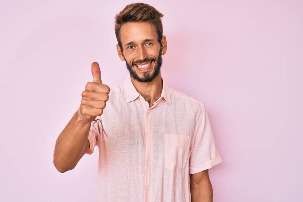Komea valkoihoinen mies, jolla on parta yllään rento vaaleanpunainen paita hymyilevä iloinen ja positiivinen, peukalo ylös tekemässä erinomaista ja hyväksyntämerkkiä  - Valokuva, kuva