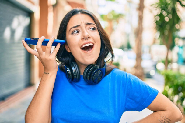 Giovane donna ispanica sorridente felice messaggio audio di ascolto utilizzando smartphone in città. - Foto, immagini