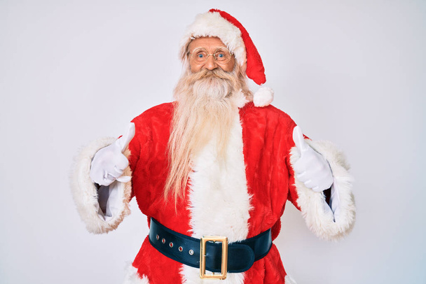 Vieil homme âgé aux cheveux gris et à la barbe longue portant le costume de Père Noël avec des bretelles approuvant faire un geste positif avec la main, les pouces levés souriants et heureux pour le succès. geste gagnant.  - Photo, image