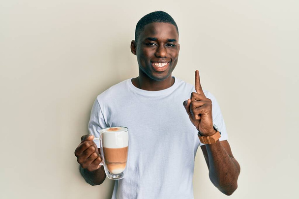 Joven hombre afroamericano sosteniendo café capuchino sonriendo con una idea o pregunta apuntando con el dedo con la cara feliz, número uno  - Foto, Imagen