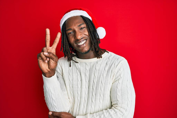 Αφροαμερικάνος με κοτσίδες που φοράει χριστουγεννιάτικο καπέλο χαμογελώντας με χαρούμενο πρόσωπο που κλείνει το μάτι στην κάμερα κάνοντας το σήμα της νίκης με τα δάχτυλα. Νούμερο δύο..  - Φωτογραφία, εικόνα