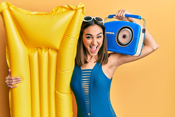 Jeune fille brune portant bikini tenant boombox et flotteur célébrant fou et étonné pour le succès avec les yeux ouverts hurlant excité.  - Photo, image
