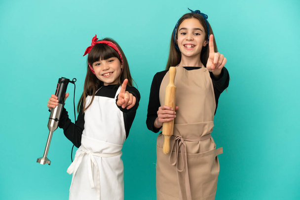 Little sisters cooking at home isolated on blue background showing and lifting a finger - Foto, Bild