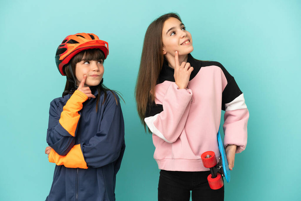 Kleine Schwestern üben Radfahren und Skater isoliert auf blauem Hintergrund denken eine Idee, während sie nach oben schauen - Foto, Bild
