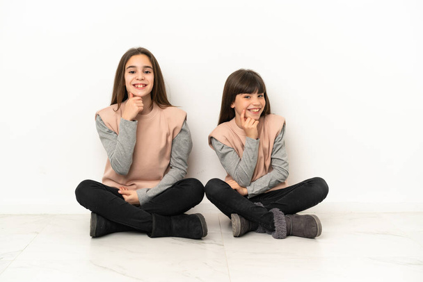 Siostry siedzące na podłodze odizolowane na białym tle uśmiechnięte słodkim wyrazem twarzy - Zdjęcie, obraz