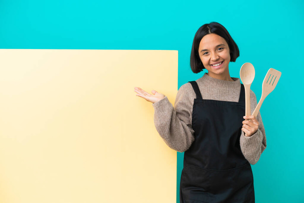 Jeune femme cuisinière métisse avec une grande pancarte isolée sur fond bleu tenant imaginaire copyspace sur la paume pour insérer une annonce et avec les pouces vers le haut - Photo, image