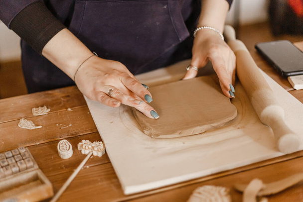 Frauenhände rollen den Ton mit einem Nudelholz auf dem Tisch in der Töpferei aus - Foto, Bild