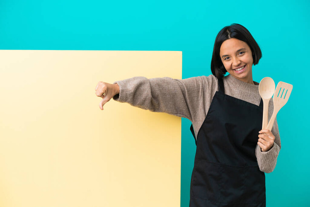 Молодая женщина-повар смешанной расы с большим плакатом на синем фоне показывает большой палец вниз с отрицательным выражением - Фото, изображение