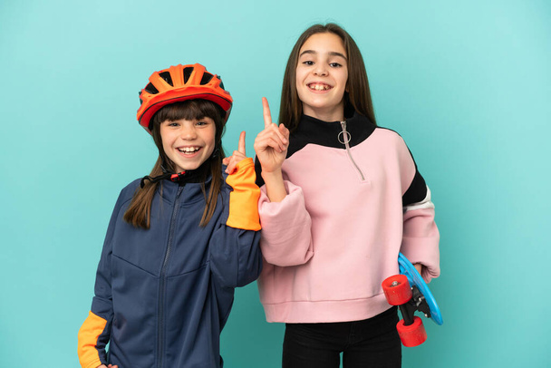 Μικρές αδελφές εξάσκηση ποδηλασία και σκέιτερ απομονώνονται σε μπλε φόντο δείχνει και να άρει ένα δάχτυλο σε ένδειξη του καλύτερου - Φωτογραφία, εικόνα