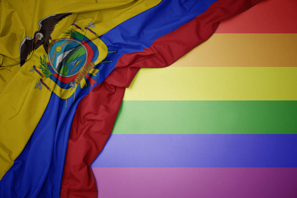 κυματίζει πολύχρωμη εθνική σημαία του Εκουαδόρ σε φόντο γκέι σημαία ουράνιο τόξο. - Φωτογραφία, εικόνα