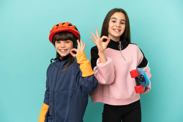 Kleine Schwestern üben Radfahren und Skater isoliert auf blauem Hintergrund zeigt ein ok Zeichen mit Fingern - Foto, Bild