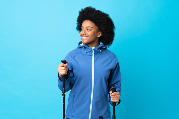 Young Africa American with backpack and trekking poles isolated on blue background looking side - Photo, Image