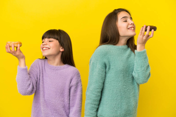 Little sisters isolated on yellow background holding donuts with happy expression - Photo, Image