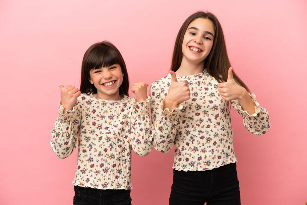 Küçük kız kardeşler pembe arka planda izole edilmiş iki elle baş parmak işareti yapıyor ve gülümsüyor. - Fotoğraf, Görsel