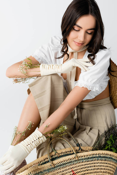 smiling woman in spring outfit and gloves with flowers sitting while posing isolated on white - Foto, imagen